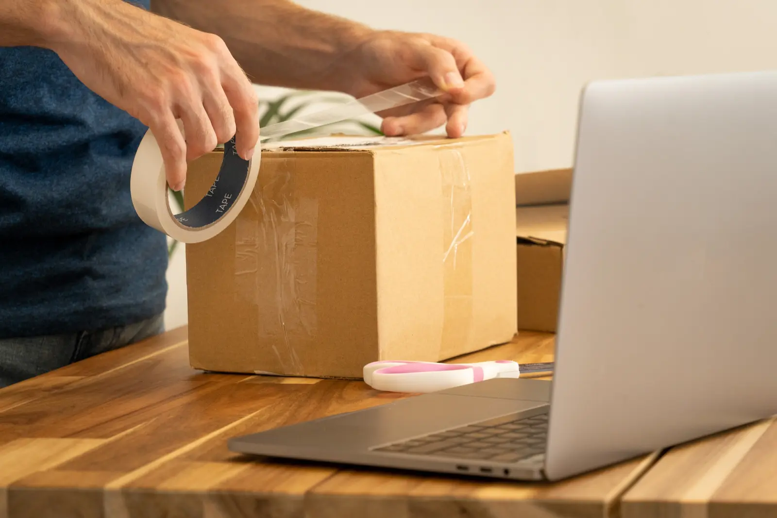 Hands sealing a cardboard parcel with tape next to a laptop